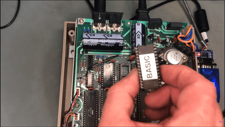 A hand holds a socketed EPROM labelled “BASIC” above the main PCB of a vintage Commodore 1541-style floppy drive, with large capacitors and a small USB-to-serial adapter visible.
